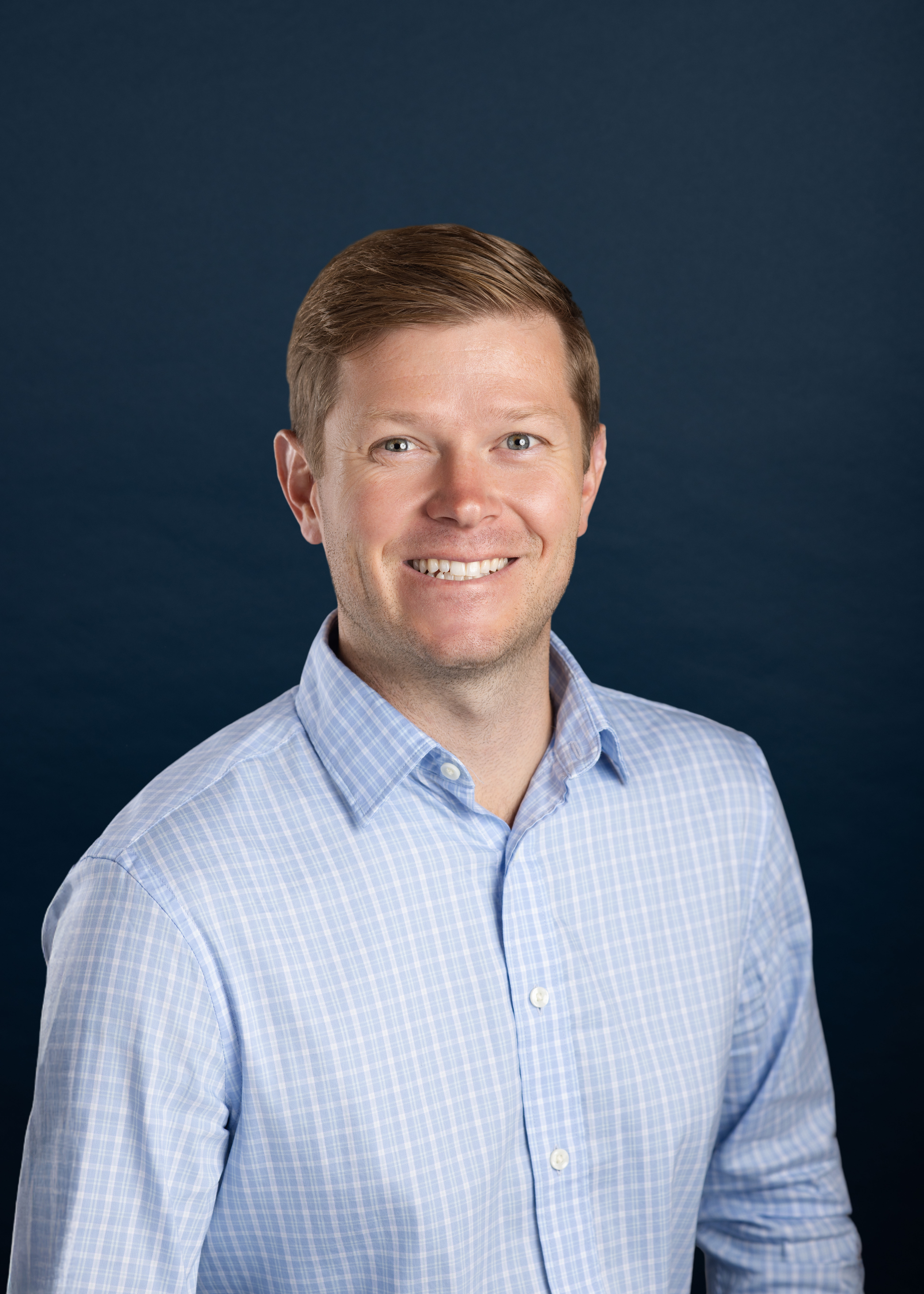Portrait of Dan Sincavage, founder of Synagentis Group, wearing a light blue checked shirt against a navy background.